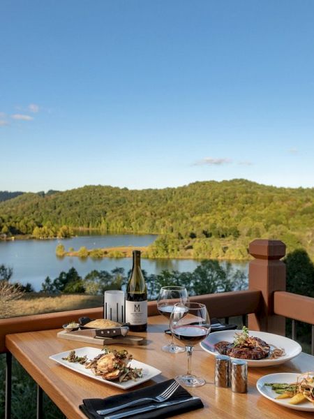 Dining setup on a deck with a lake view, wine, and food under a clear sky.