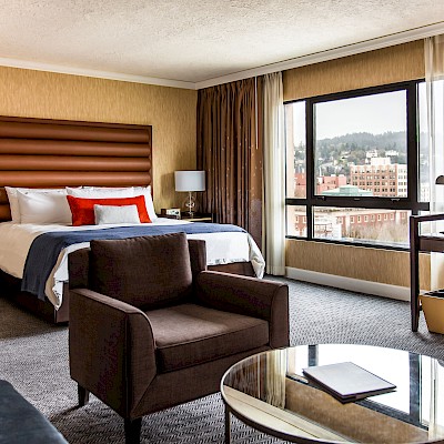A modern hotel room with a bed, armchair, desk, and a view of the city through a large window. The decor is stylish and inviting.