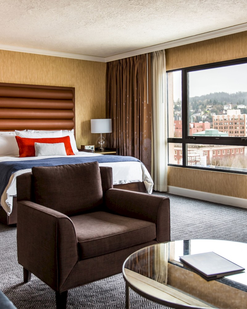 A modern hotel room with a bed, armchair, desk, and a view of the city through a large window. The decor is stylish and inviting.