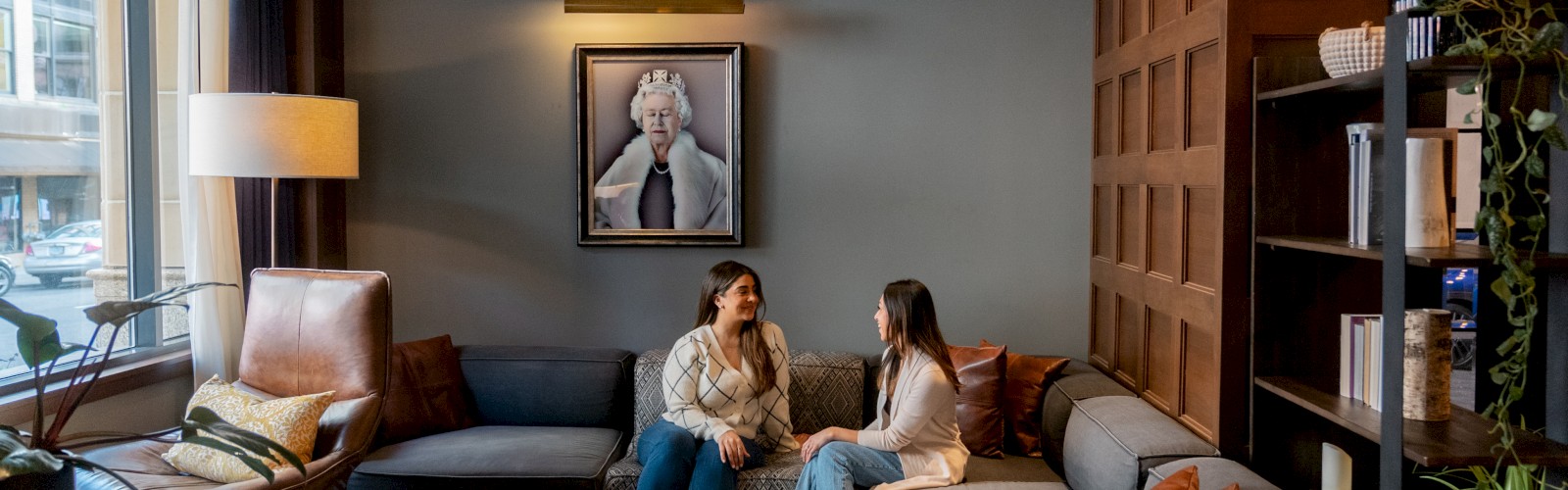 Two people sit on a couch in a cozy living room with a large window, plants, a lamp, and a framed photo on the wall behind them.
