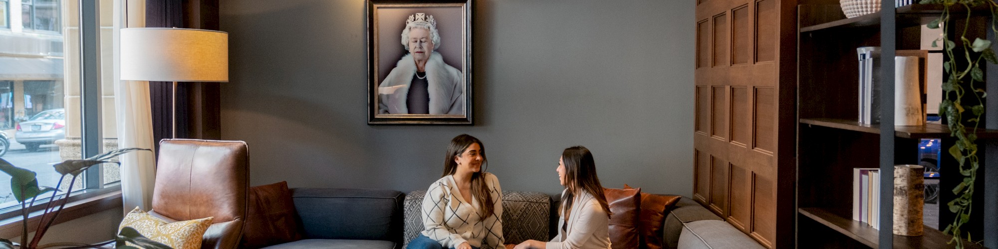 Two people sit on a couch in a cozy living room with a large window, plants, a lamp, and a framed photo on the wall behind them.