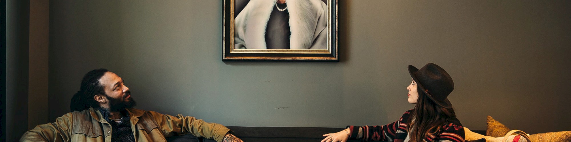 Two people sit on a couch, facing each other, with a portrait of a queen between them on the wall, and bags beside the woman.