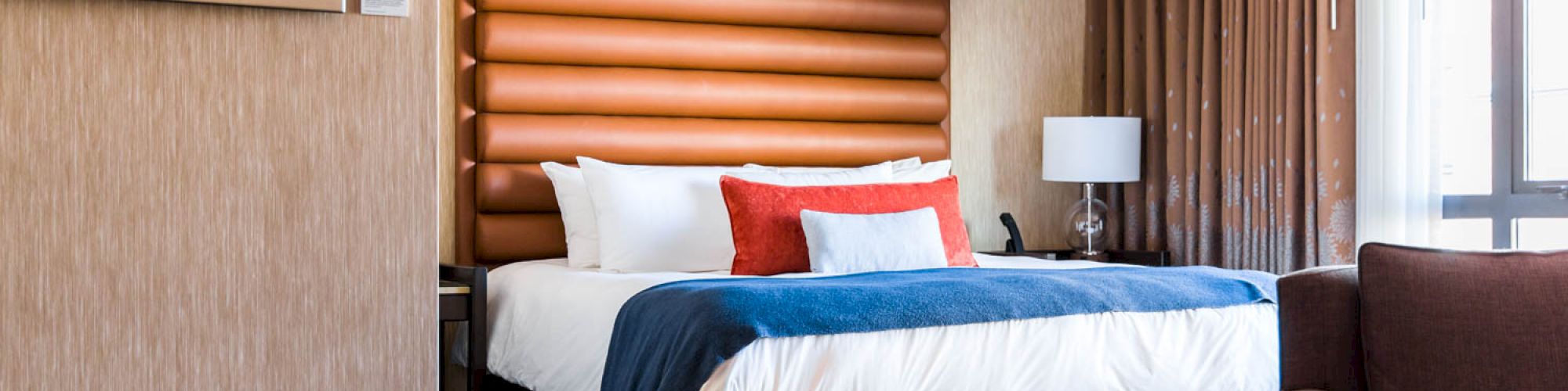 A modern hotel room features a large bed with blue and red accents, a window with brown curtains, a lamp, and a comfortable seating area.