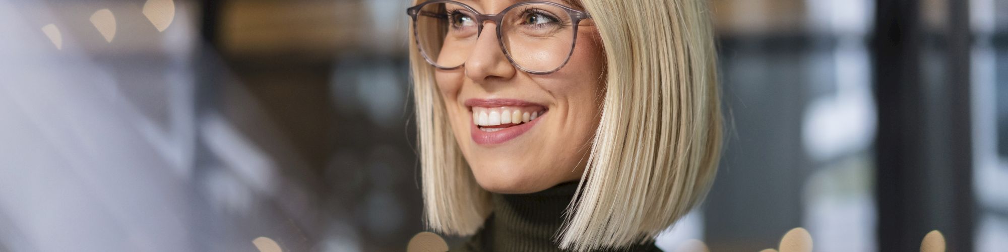 A person with blonde hair and glasses is smiling, wearing a dark green turtleneck sweater. The background is blurry with round lights and window reflections.