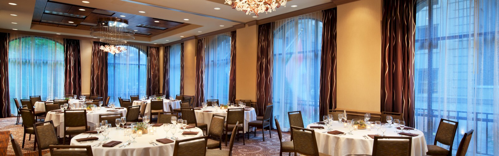 A well-lit banquet room with round tables arranged for a formal event, lavish chandeliers, patterned carpet, and large windows with curtains.
