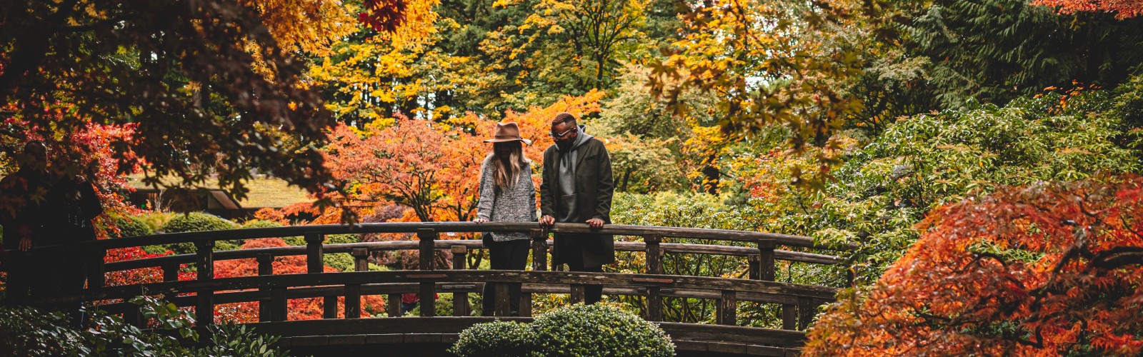 A couple stands on a wooden bridge in a lush, colorful autumn garden with vibrant orange, red, and yellow foliage surrounding them.