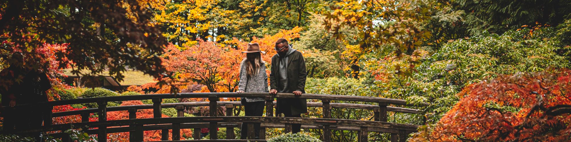 A couple stands on a wooden bridge in a lush, colorful autumn garden with vibrant orange, red, and yellow foliage surrounding them.