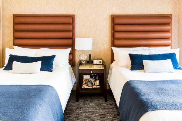 The image shows a hotel room with two adjacent beds, each with blue blankets and headboards. A nightstand with a lamp and phone is between them.