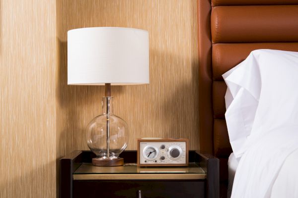 The image shows a bedside table with a glass lamp and a vintage radio beside a bed with white sheets in a well-lit room.