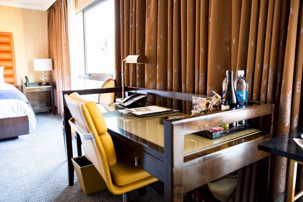 A hotel room features a desk with a chair, lamp, telephone, bottled water, and snacks, next to a window with curtains and a bed in the background.
