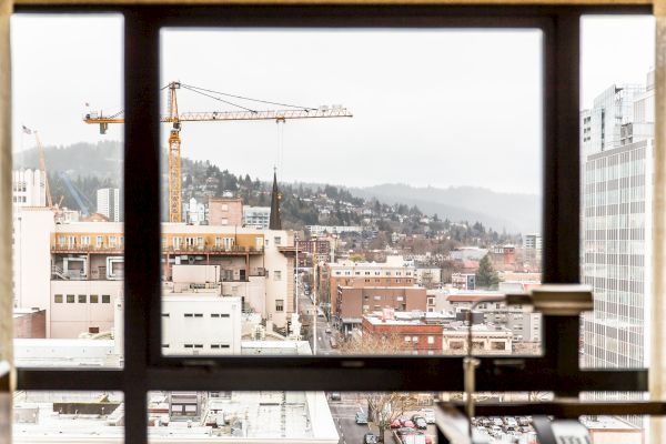 A cityscape is viewed through a window, featuring buildings, a crane, and hills in the background under a cloudy sky, ending the sentence.