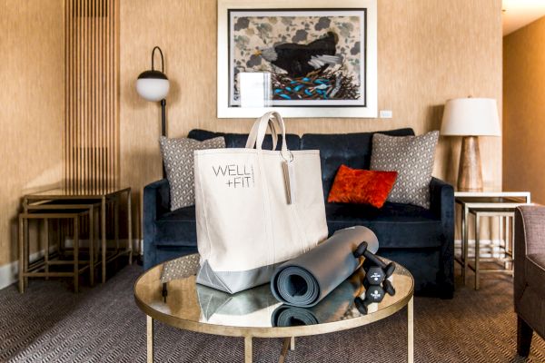 A living room with a blue sofa, decorative cushions, a framed picture, a round glass table with a wellness bag, yoga mat, and dumbbells on it.