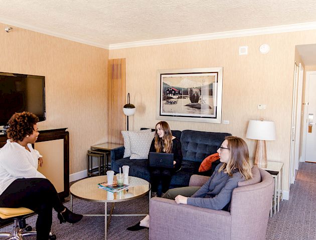 Three people are having a conversation in a cozy living room with a TV, sofa, chair, and a coffee table.