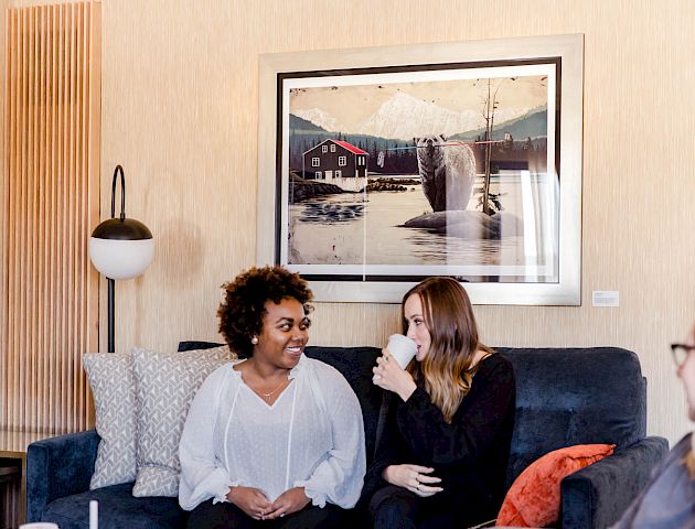 Three women are indoors, sitting on a couch and talking. Behind them is a framed picture of a whale in water. Ending the sentence.