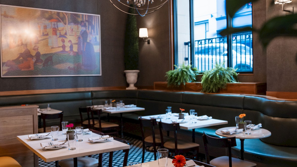 This image shows a cozy, elegant restaurant with chandeliers, decorated tables, and a large framed picture on the wall, viewed through some foliage.