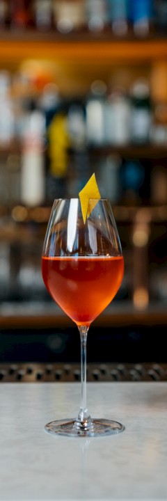 A fancy cocktail in a stemmed glass, topped with a red/orange gradient, set against a blurred bar backdrop, bright lighting.