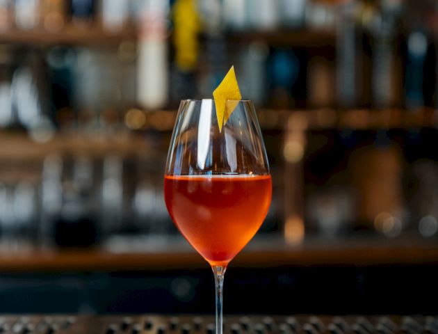 A glass of red-colored cocktail garnished with a twist of lemon peel is placed on a bar counter with a blurred background of bottles.