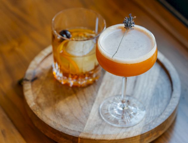 The image shows two cocktails on a round wooden tray: a brown drink in a rocks glass with an ice sphere, and an orange cocktail in a coupe glass.