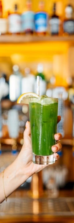 A hand holds a tall glass of green juice garnished with a lime slice, with a bar displaying numerous bottles in the background.