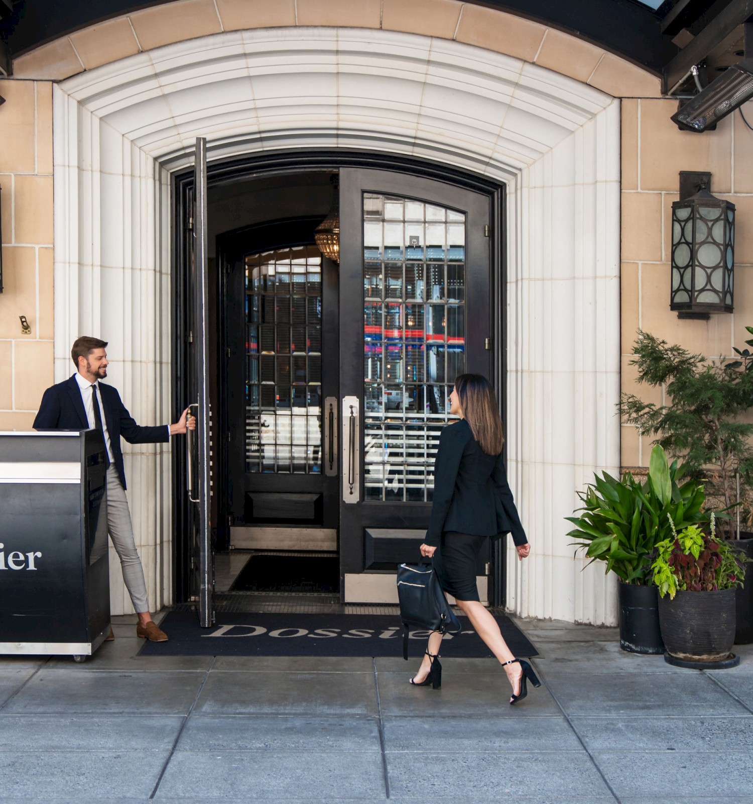 A person is entering a building named Dossier while another person holds the door open. The entrance is stylish with plants and decor.