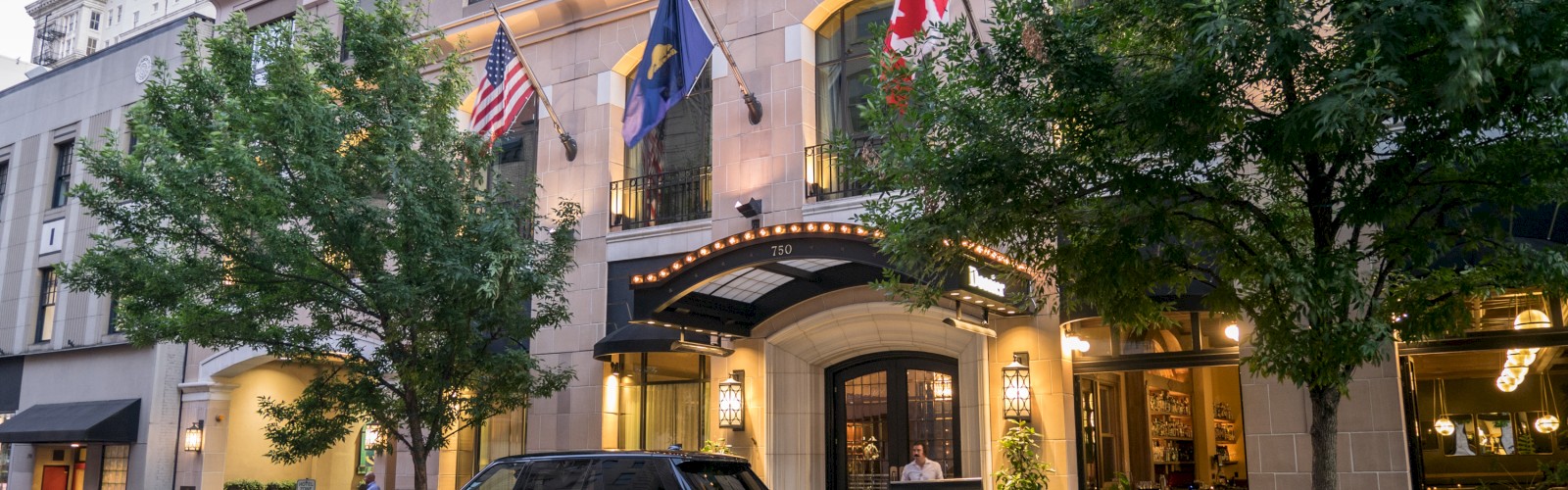 The image shows a street view of a building with flags, trees, and a parked car in front of a hotel entrance.
