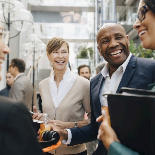 A group of four professionals are engaging in a lively conversation at a social gathering or networking event, smiling and holding documents.