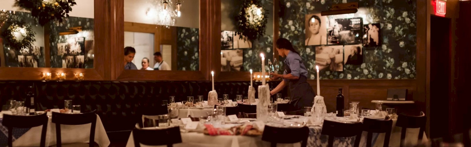 A dimly lit, elegant restaurant with chandeliers, prepared tables, floral wallpaper, and a server arranging items, setting a warm ambiance.