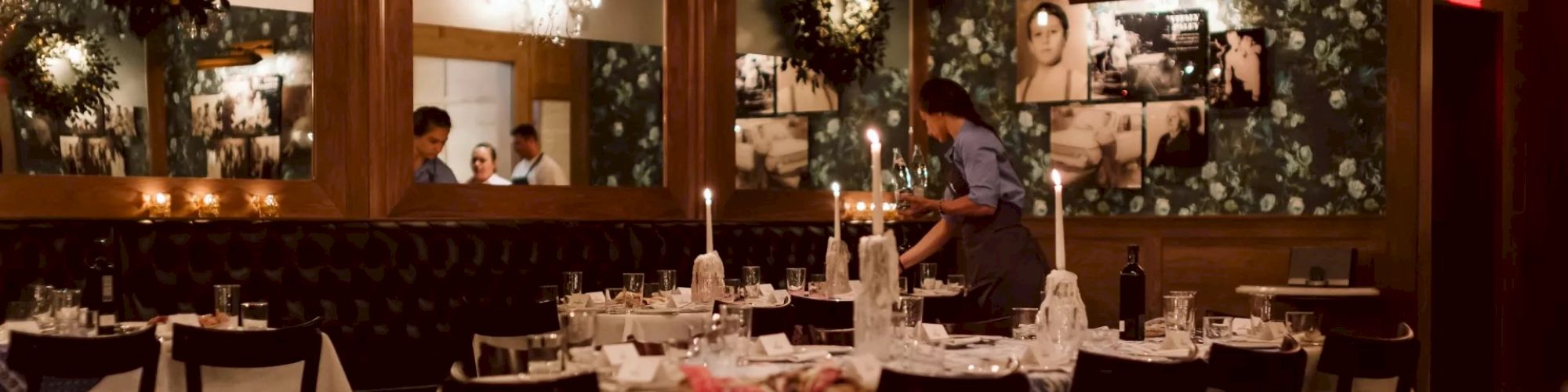 A dimly lit, elegant restaurant with chandeliers, prepared tables, floral wallpaper, and a server arranging items, setting a warm ambiance.