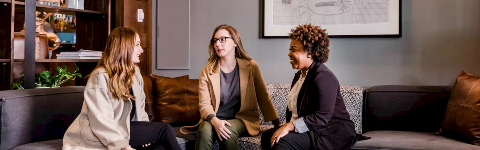 Three women are sitting on a gray couch, engaged in conversation, with a coffee table and a book in front of them. A framed drawing hangs on the wall.