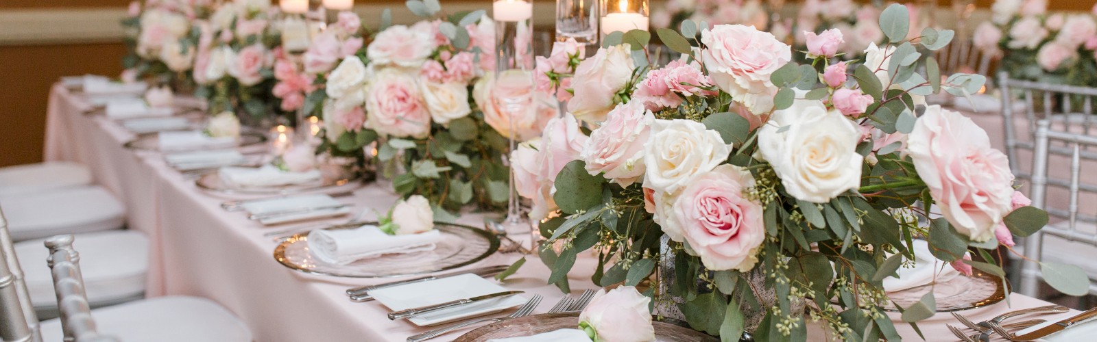 A beautifully set dinner table with elegant floral centerpieces, white plates, cutlery, and lit candles in a warmly lit dining room.