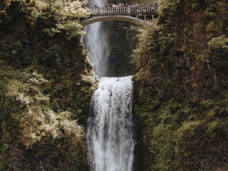 The image shows a scenic waterfall with two cascades and a bridge in between, surrounded by lush greenery and steep rock formations.