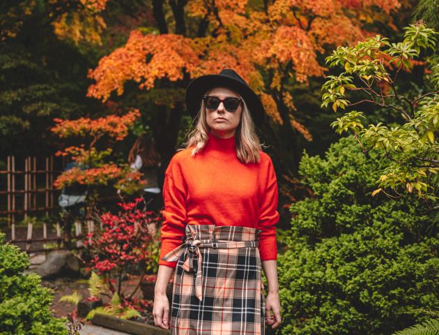 A woman in a red sweater and plaid skirt stands outdoors in a lush garden with vibrant autumn foliage, wearing sunglasses and a black hat.