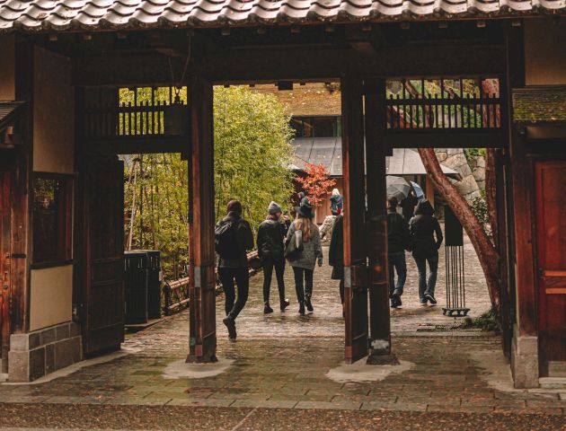 A traditional wooden gate leads to a garden with people in the background, set in autumn with vibrant orange leaves overhead.