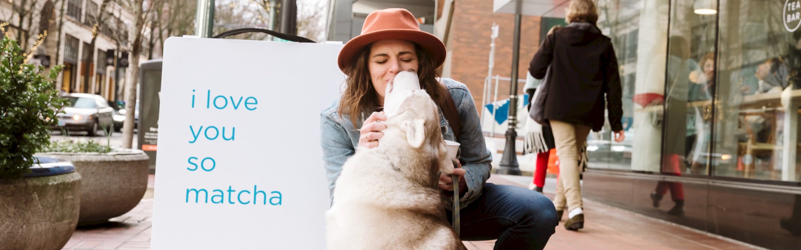 A woman in a hat crouches to kiss a dog, near a sign that reads 