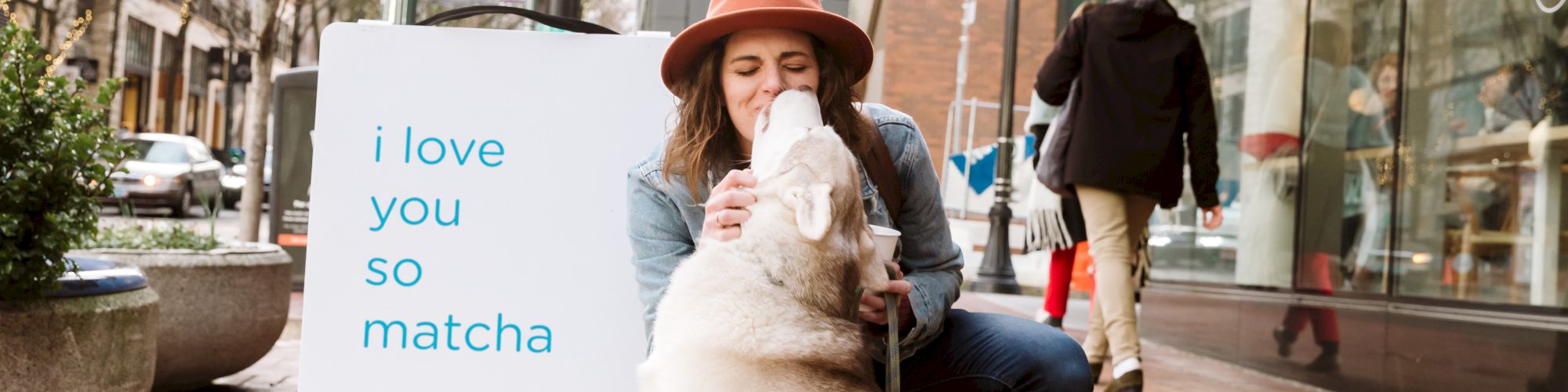 A woman in a hat crouches to kiss a dog, near a sign that reads 