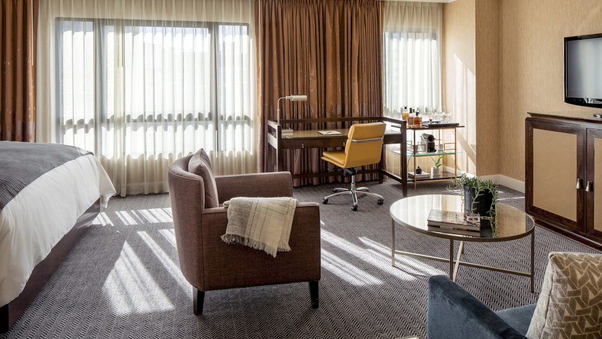 A spacious hotel room with a bed, armchair, desk, TV, and large windows with curtains, filled with cozy natural sunlight.