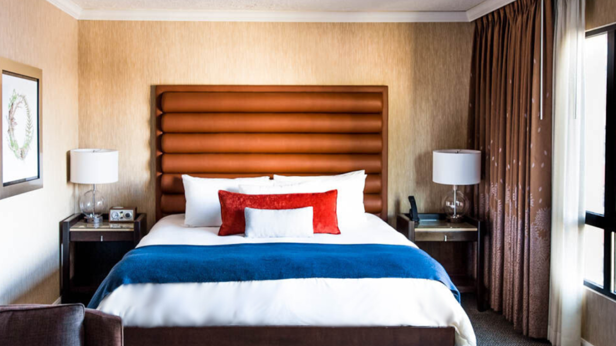 A well-decorated hotel room with a large bed, vibrant pillows, lamps on nightstands, armchair, and a window view of a red brick building.