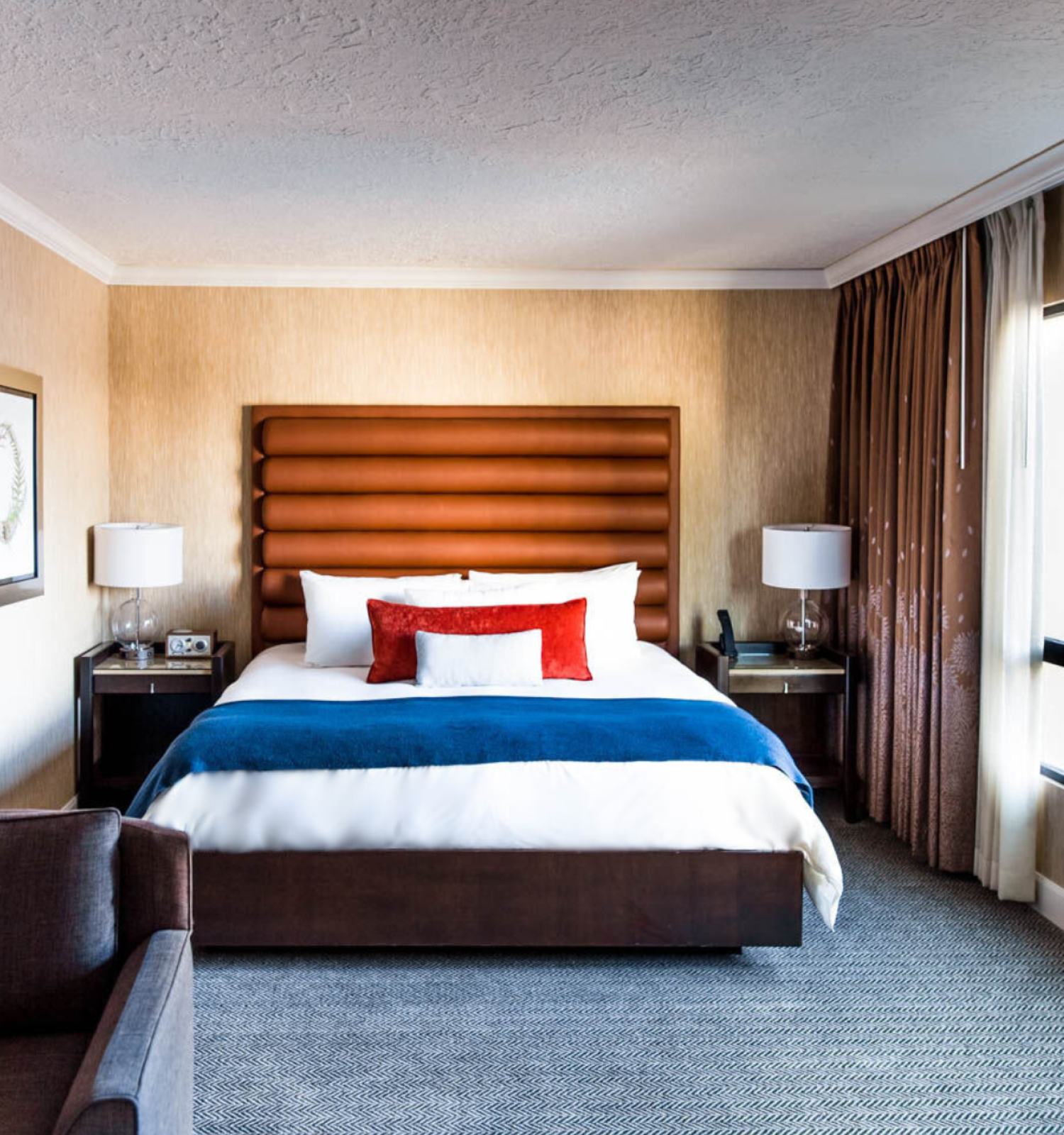 A well-decorated hotel room with a large bed, vibrant pillows, lamps on nightstands, armchair, and a window view of a red brick building.