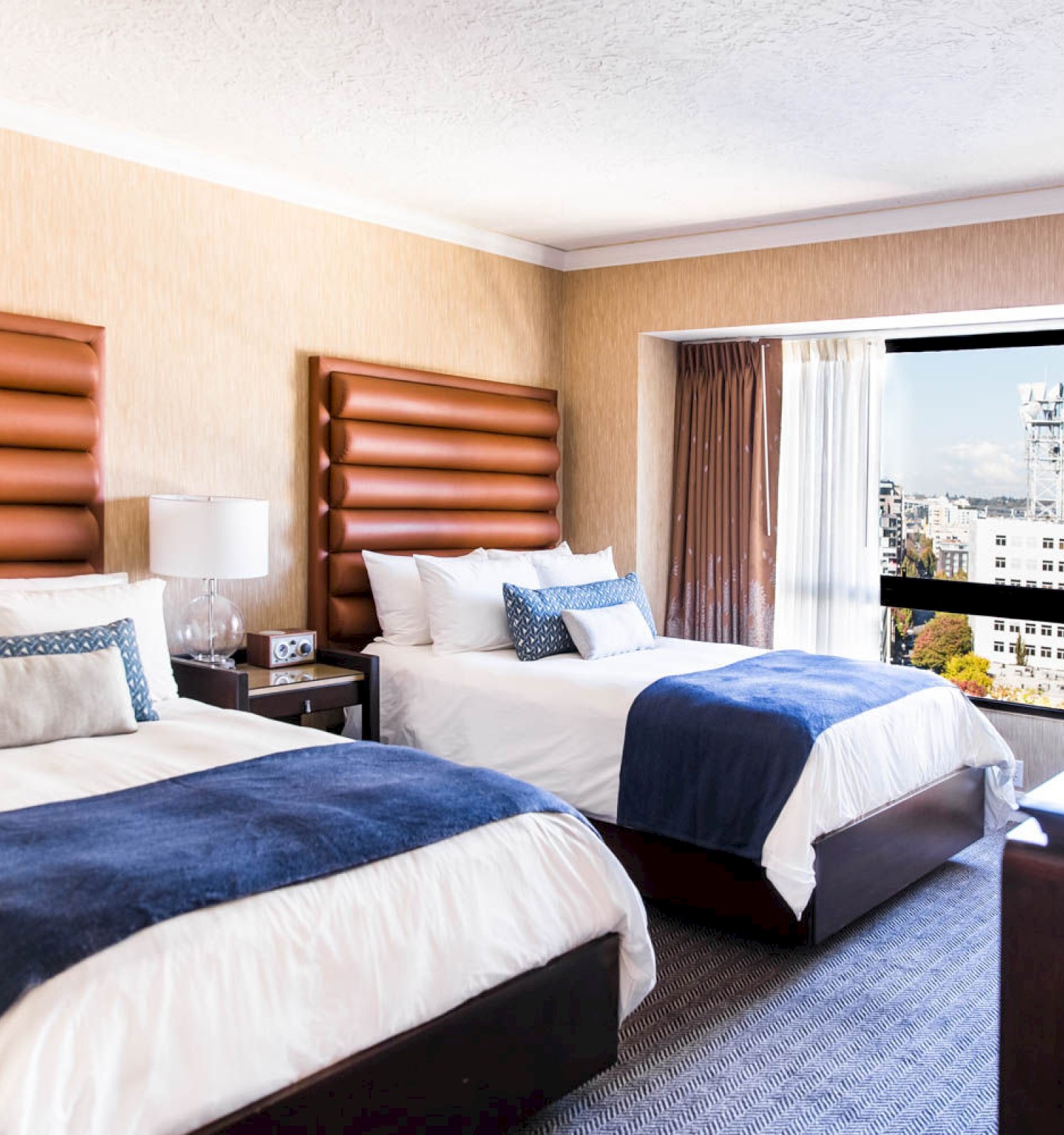 The image shows a hotel room with two beds, plush headboards, a window view, bedside table, and a lamp, creating a cozy atmosphere.