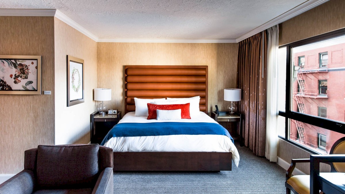 A cozy hotel room featuring a bed with white linens, red and blue accents, lamps, artwork, and a view of buildings outside the window.