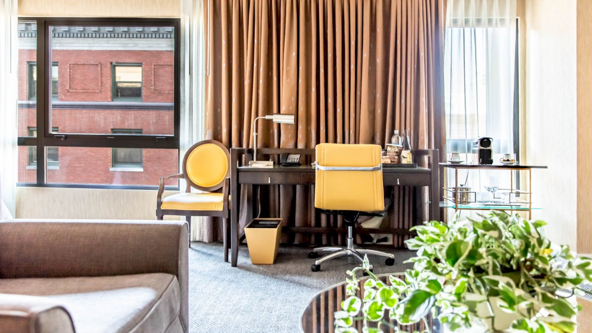 A well-lit hotel room with a desk, chairs, sofa, and a view of the outside building.