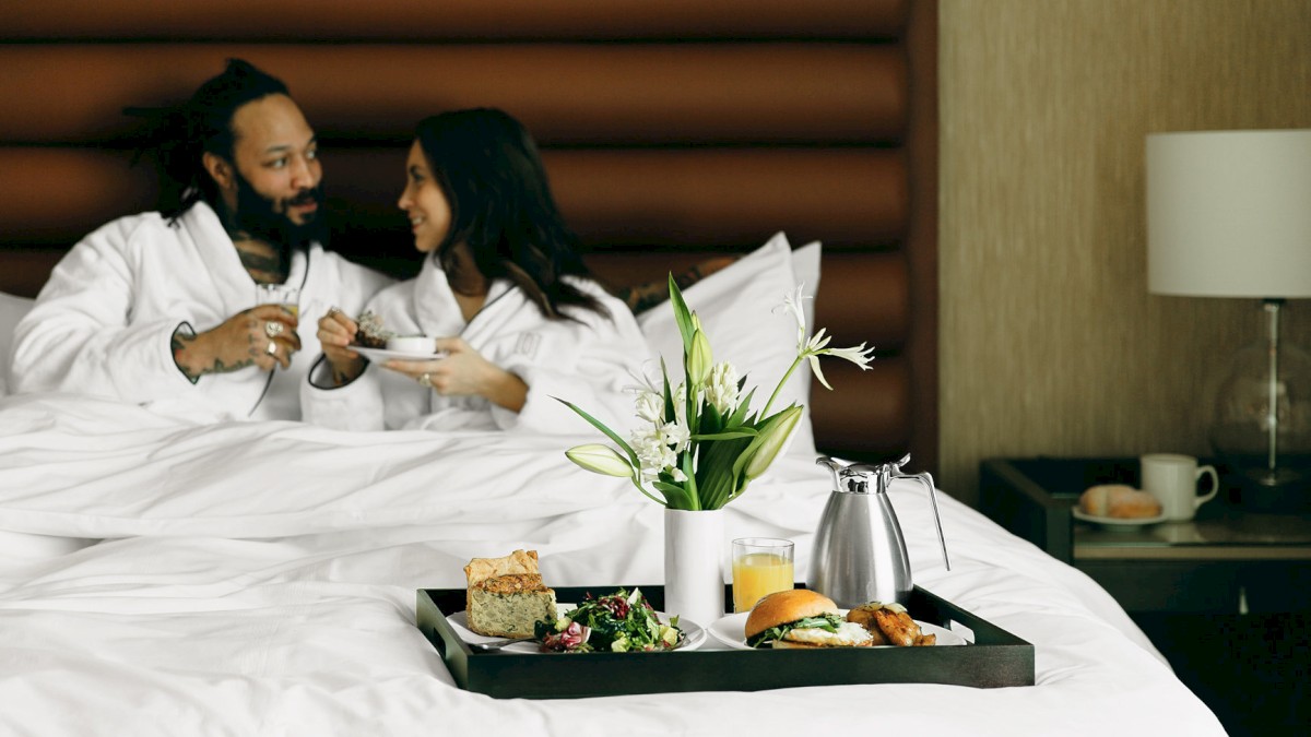 A couple in robes enjoys breakfast in bed with pastries, juice, and flowers on a tray, creating a cozy ambiance.