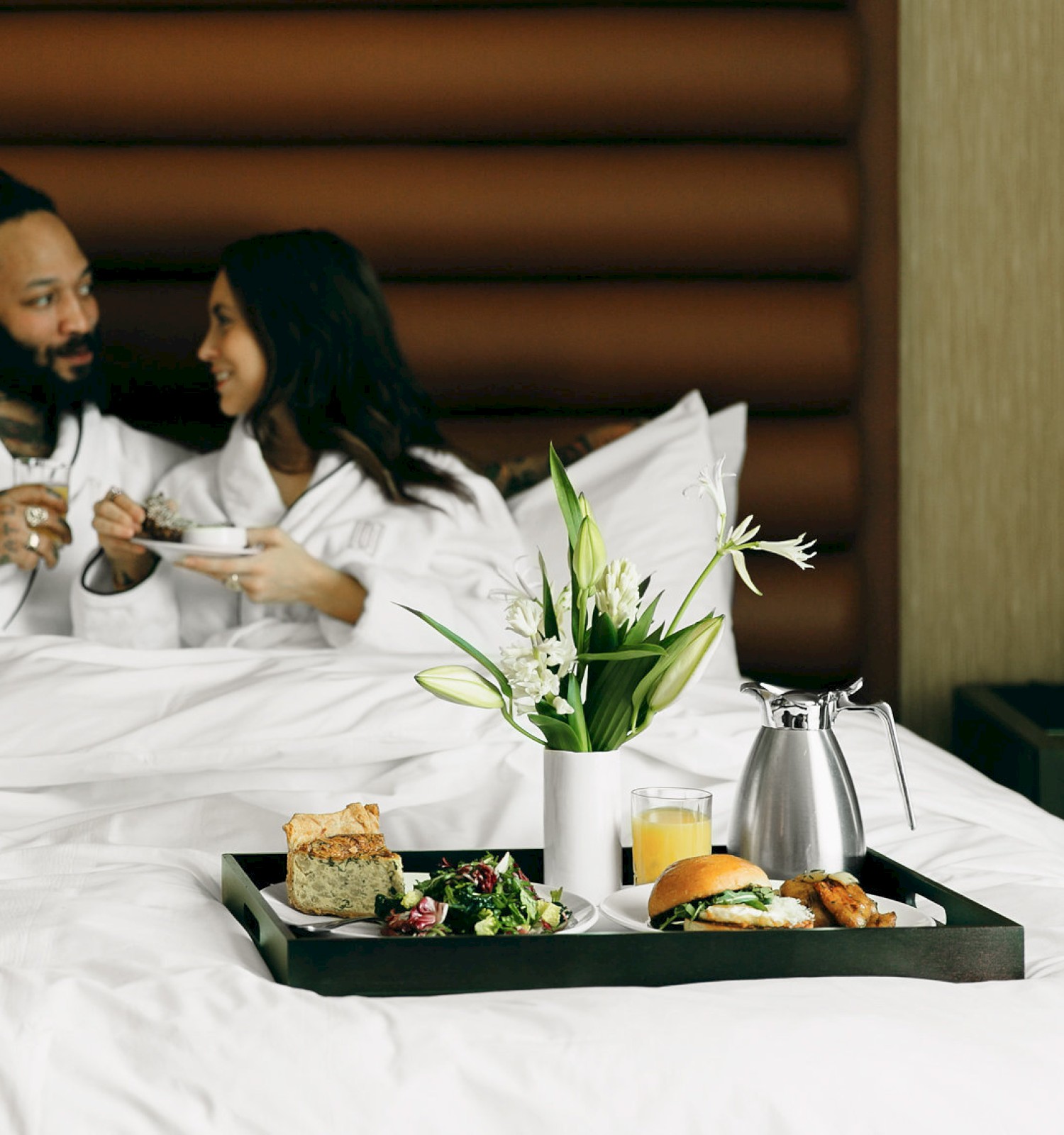 A couple in robes enjoys breakfast in bed with pastries, juice, and flowers on a tray, creating a cozy ambiance.