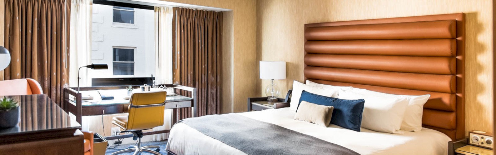 A hotel room with a large bed, desk, chair, lamp, and window with curtains, offering a modern and cozy environment for guests.
