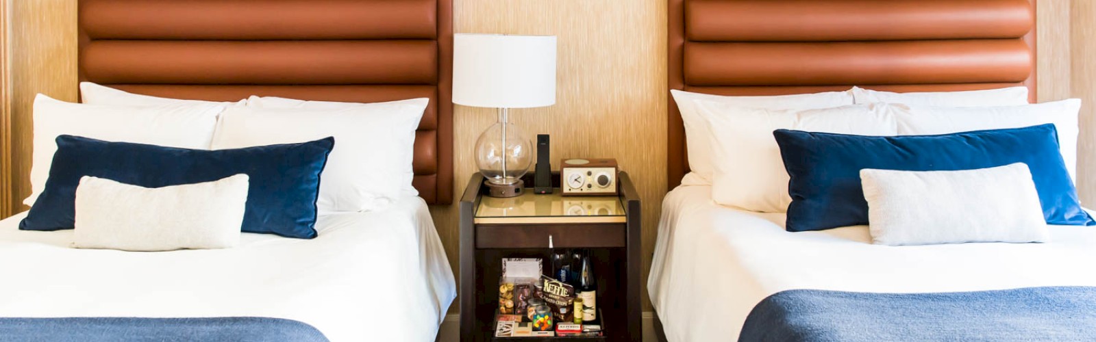 The image shows a hotel room with two beds, each with blue bed runners and white bedding; a nightstand with a lamp and amenities is in the center.