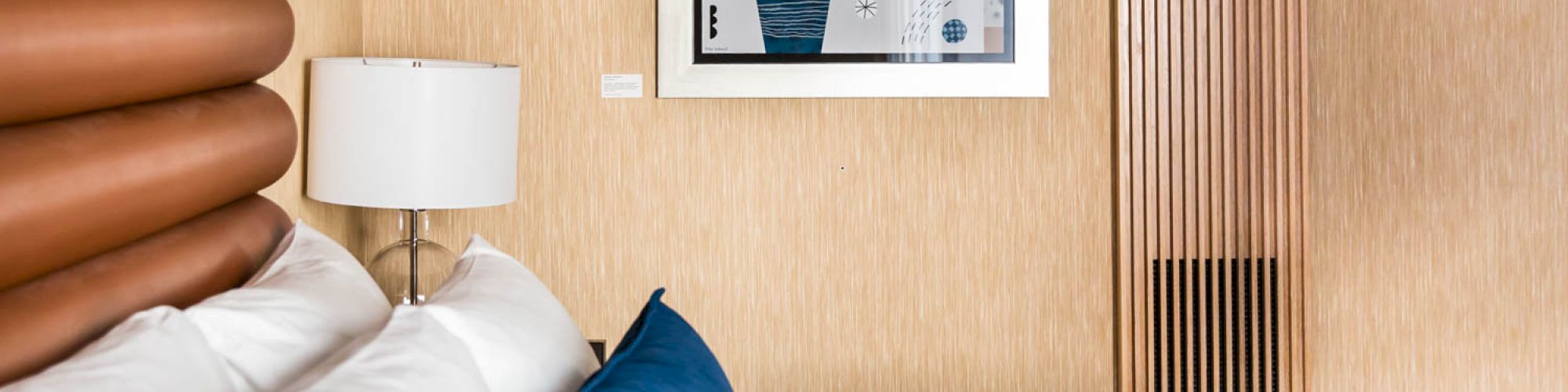 The image shows a modern hotel room with a neatly made bed, blue and white pillows, a lamp, and a framed artwork of a plant on the wall.