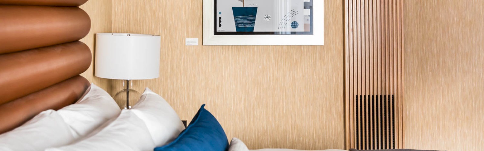 The image shows a neatly arranged bed with white and blue pillows, a wall lamp, and framed artwork on the wall. The room is tastefully decorated.