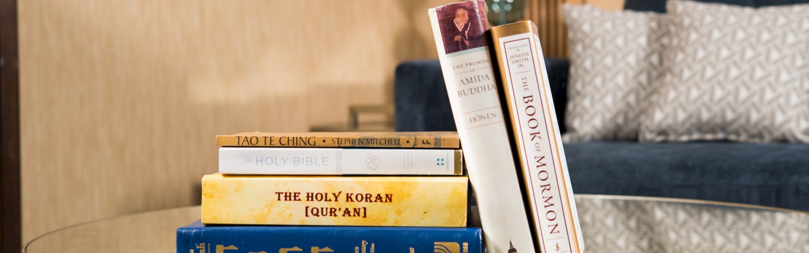 A stack of religious books, including The Holy Bible, The Holy Koran (Qur’an), and The Book of Mormon, on a glass table in a living room setting.