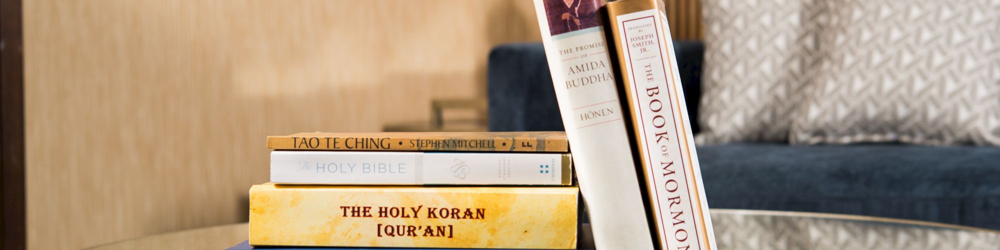 A stack of religious books, including The Holy Bible, The Holy Koran (Qur’an), and The Book of Mormon, on a glass table in a living room setting.