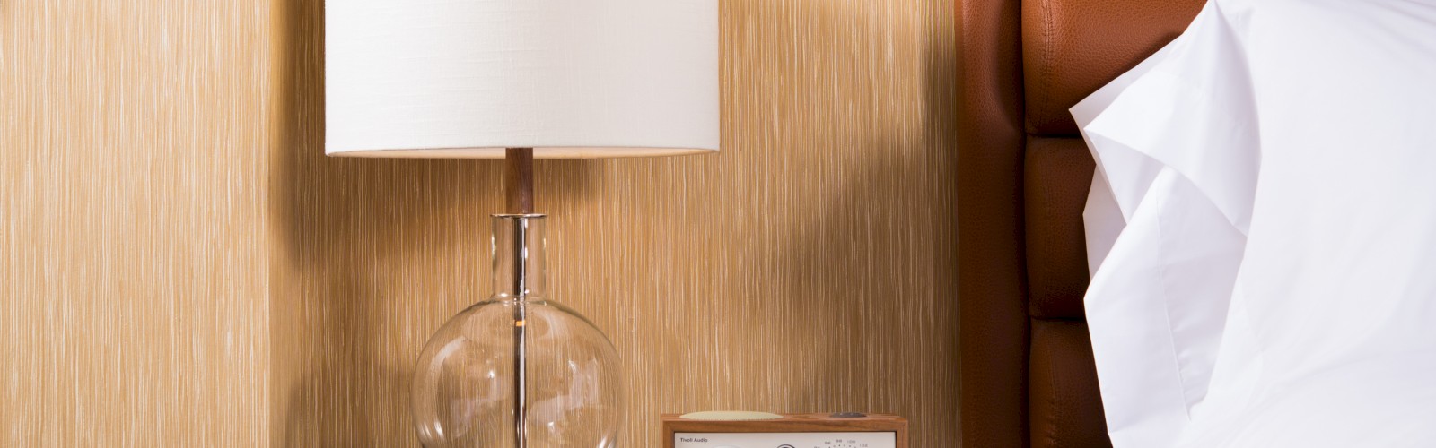 A bedside table with a lamp featuring a clear glass base, a retro-style radio, and a portion of a bed with white linens is shown.